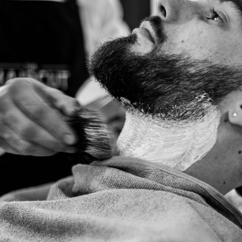 Barbeiro aplicando espuma e preparando a barba de um cliente para o barbear.