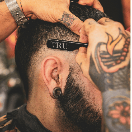 Barbeiro realizando corte de cabelo com precisão, utilizando uma navalha com a inscrição "TRU".